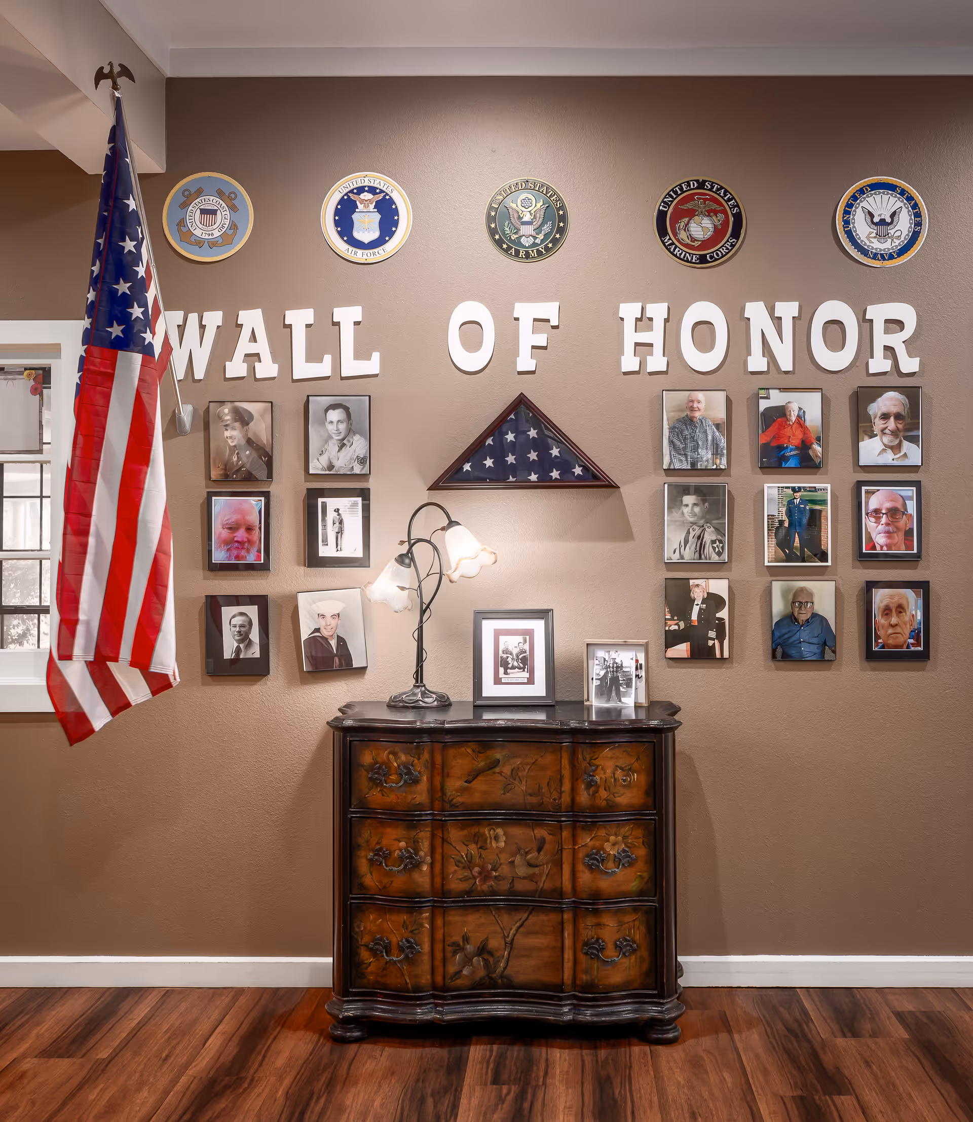A Wall of Honor display featuring framed photographs of veterans, a folded American flag in a triangular case, and military branch emblems above. An American flag stands to the left, and a decorative wooden chest with a lamp and additional framed photos is below the display on a wooden floor.