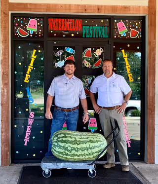 Two men standing outside a building entrance decorated with colorful watermelon and candy-themed stickers and the words 'Watermelon Festival'. In front of them is a large watermelon placed on a wheeled cart.