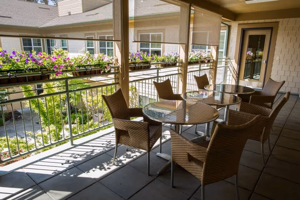 Outdoor balcony area with three round glass-top tables and six wicker chairs. Flower boxes with purple and white flowers hang on the railing. The balcony overlooks a courtyard with greenery and windows of the building are visible in the background.