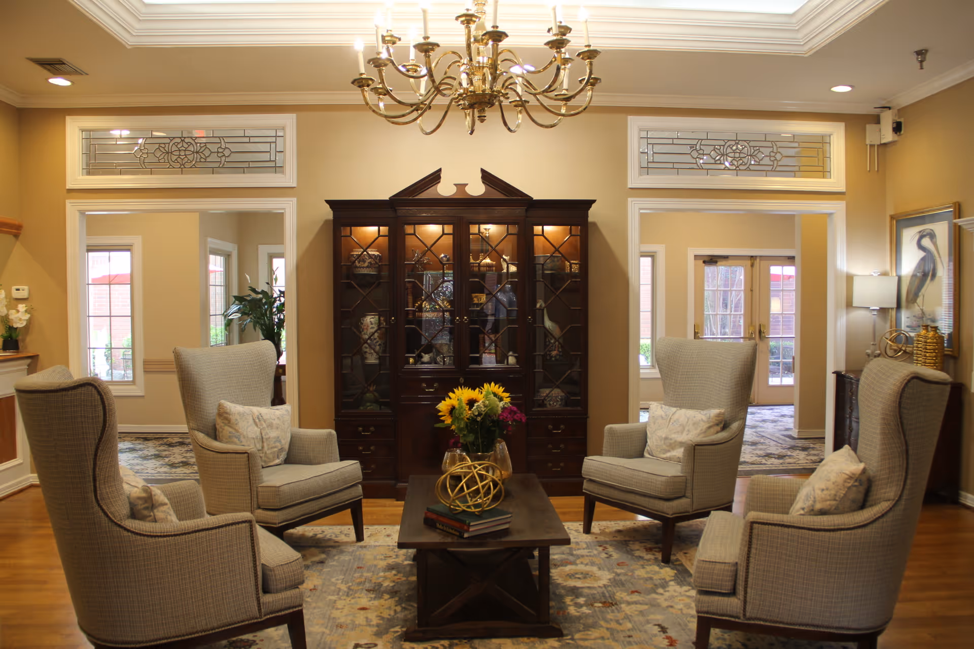 A cozy living room area with four upholstered armchairs arranged around a wooden coffee table that holds a vase of sunflowers and decorative objects. Behind the seating area is a dark wooden cabinet with glass doors displaying various decorative items. The room features beige walls, wooden flooring, a patterned rug, and a brass chandelier hanging from the ceiling. There are windows and doorways leading to other parts of the facility, with natural light coming in.