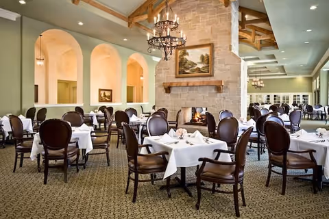 A spacious dining room with multiple round tables covered with white tablecloths and set with napkins and small floral centerpieces. The room features a large stone fireplace with a painting above it, arched wall openings, and elegant chandeliers hanging from a high ceiling with exposed wooden beams.