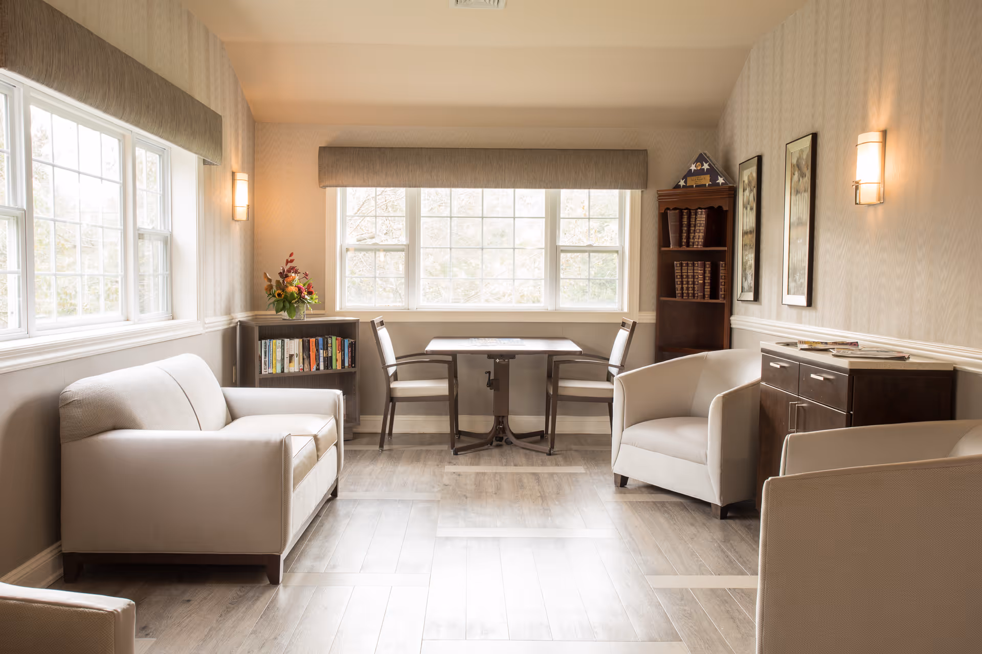 A bright and cozy sitting area with large windows letting in natural light. The room features a beige sofa, two armchairs, a small table with two chairs by the window, a bookshelf with books and a flower arrangement, and a wooden cabinet with framed artwork on the wall.