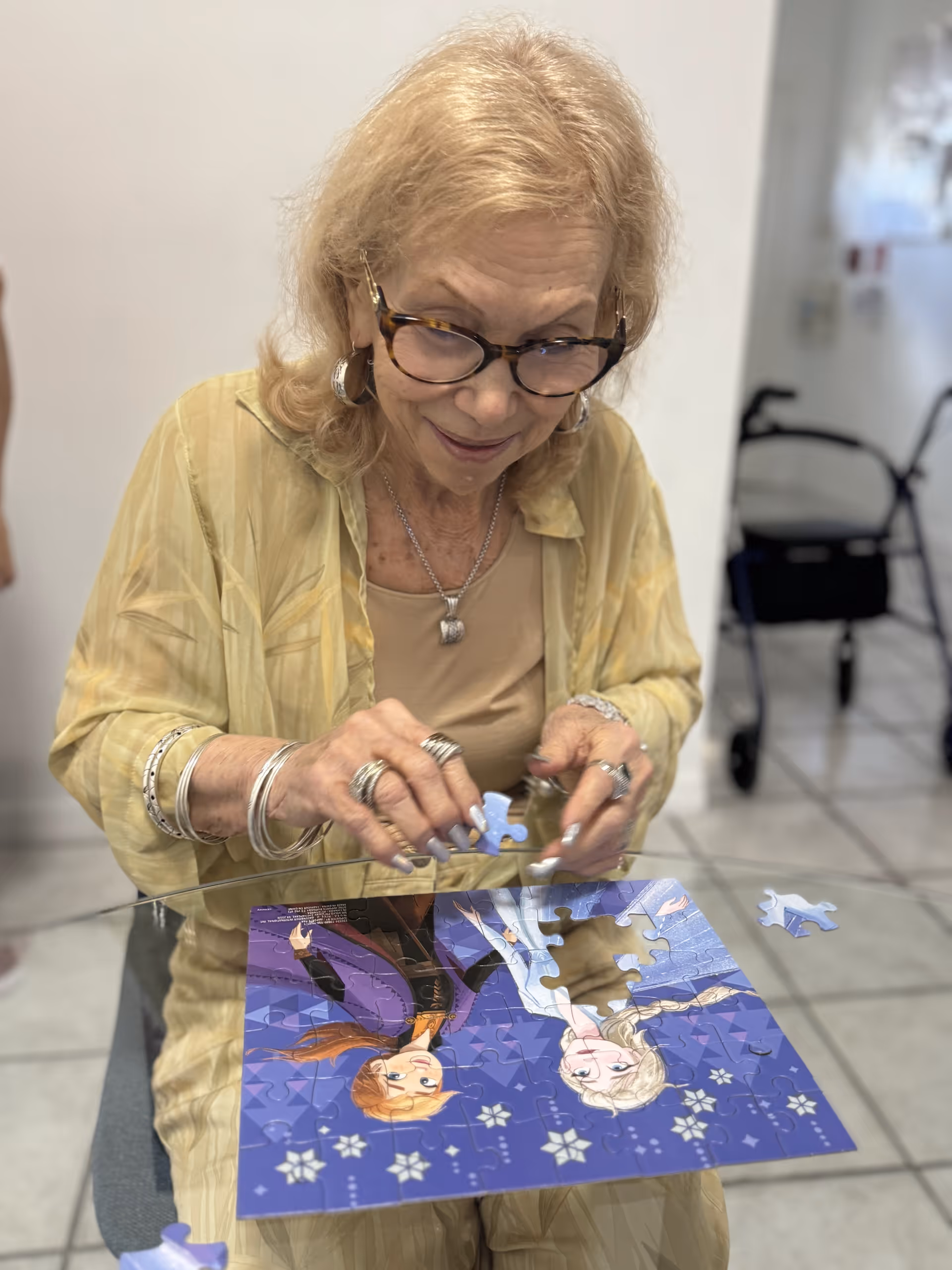 An elderly woman with glasses and light-colored hair is sitting at a glass table, assembling a jigsaw puzzle featuring characters from the movie Frozen. She is wearing a yellow shirt and multiple rings and bracelets. In the background, a walker is visible in a tiled room.