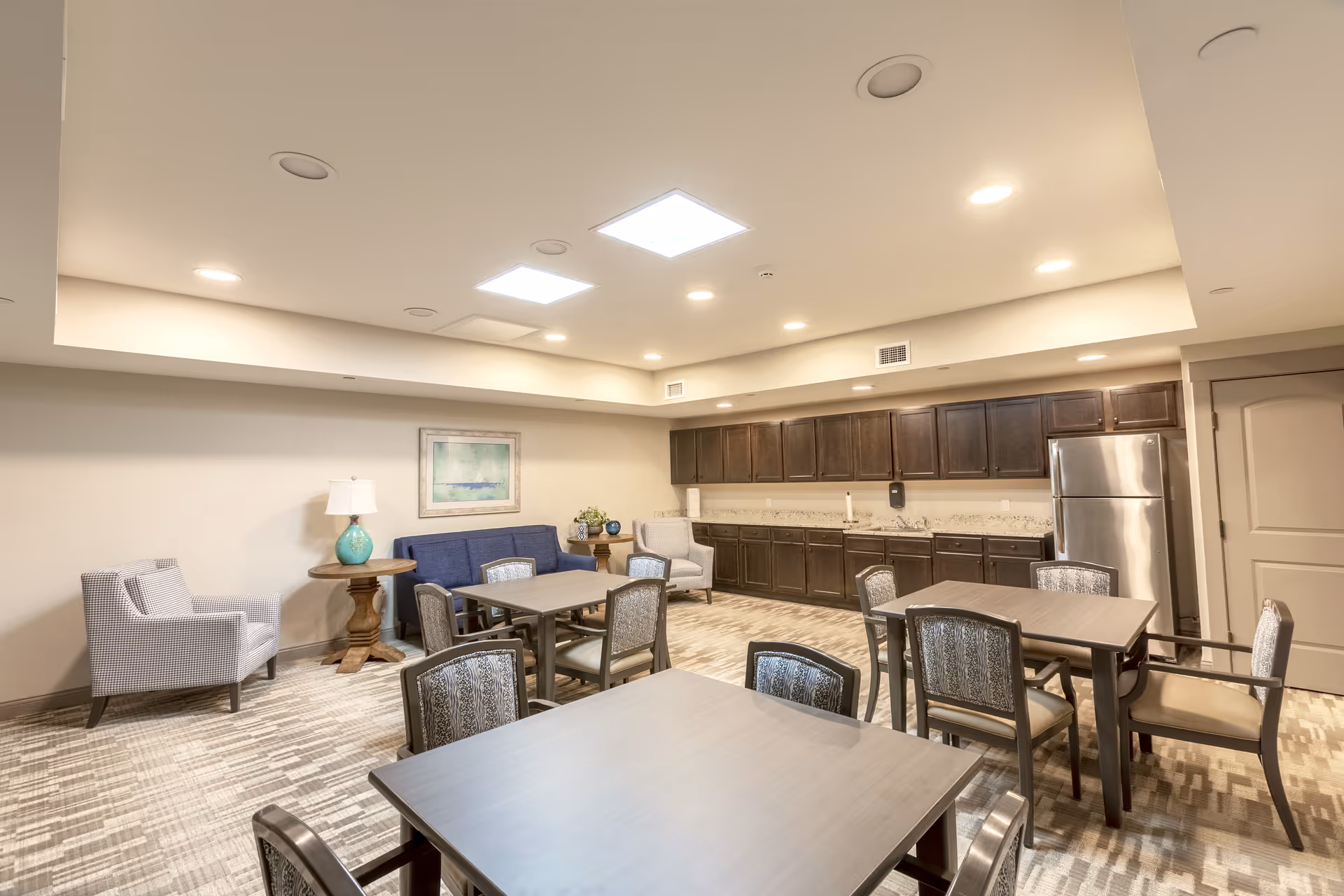 A well-lit common room with several tables and chairs arranged for seating. The room features a kitchenette area with dark wood cabinets, a stainless steel refrigerator, and a countertop. There are two armchairs and a small round table with a lamp and decorative items against one wall, along with a framed artwork. The ceiling has recessed lighting and skylights.