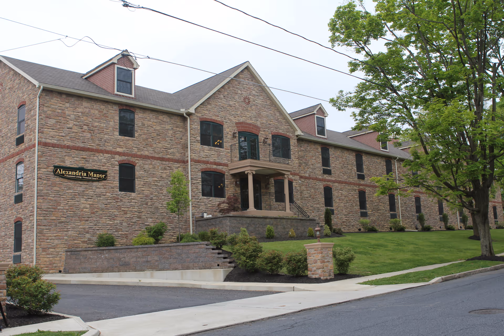 Exterior view of Alexandria Manor, a large stone building with multiple windows, a small balcony above the entrance, and a well-maintained lawn with shrubs and trees in front.