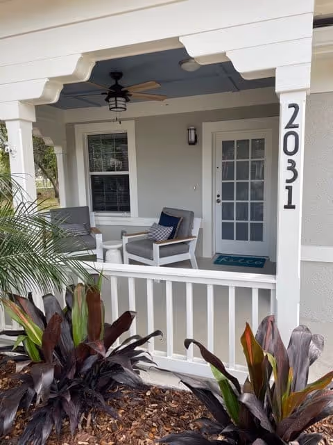 Front porch of a senior living unit with two chairs, a small table, a ceiling fan, house number 2031, and plants in the foreground.