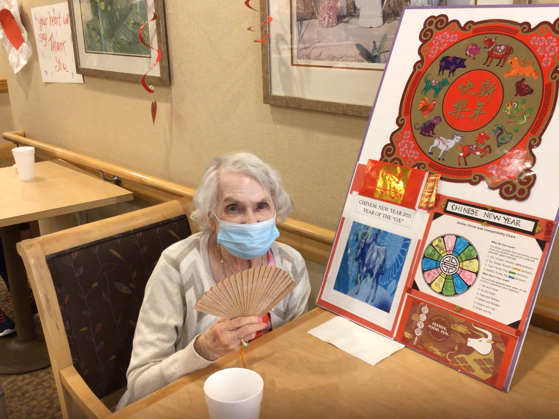 An elderly woman wearing a face mask sits at a table in a senior living facility. She is holding a wooden hand fan and smiling. On the table next to her is a colorful display board about Chinese New Year, featuring zodiac animals and a compatibility chart. The room has framed artwork on the walls and a handrail along the wall behind her.