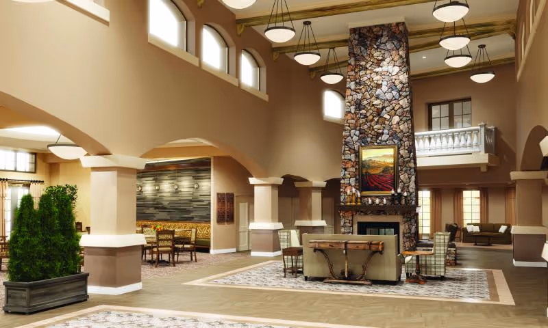Spacious senior living facility common area with high ceilings, large stone fireplace, multiple seating arrangements including armchairs and sofas, decorative plants, and warm beige walls accented by wooden beams and arches.