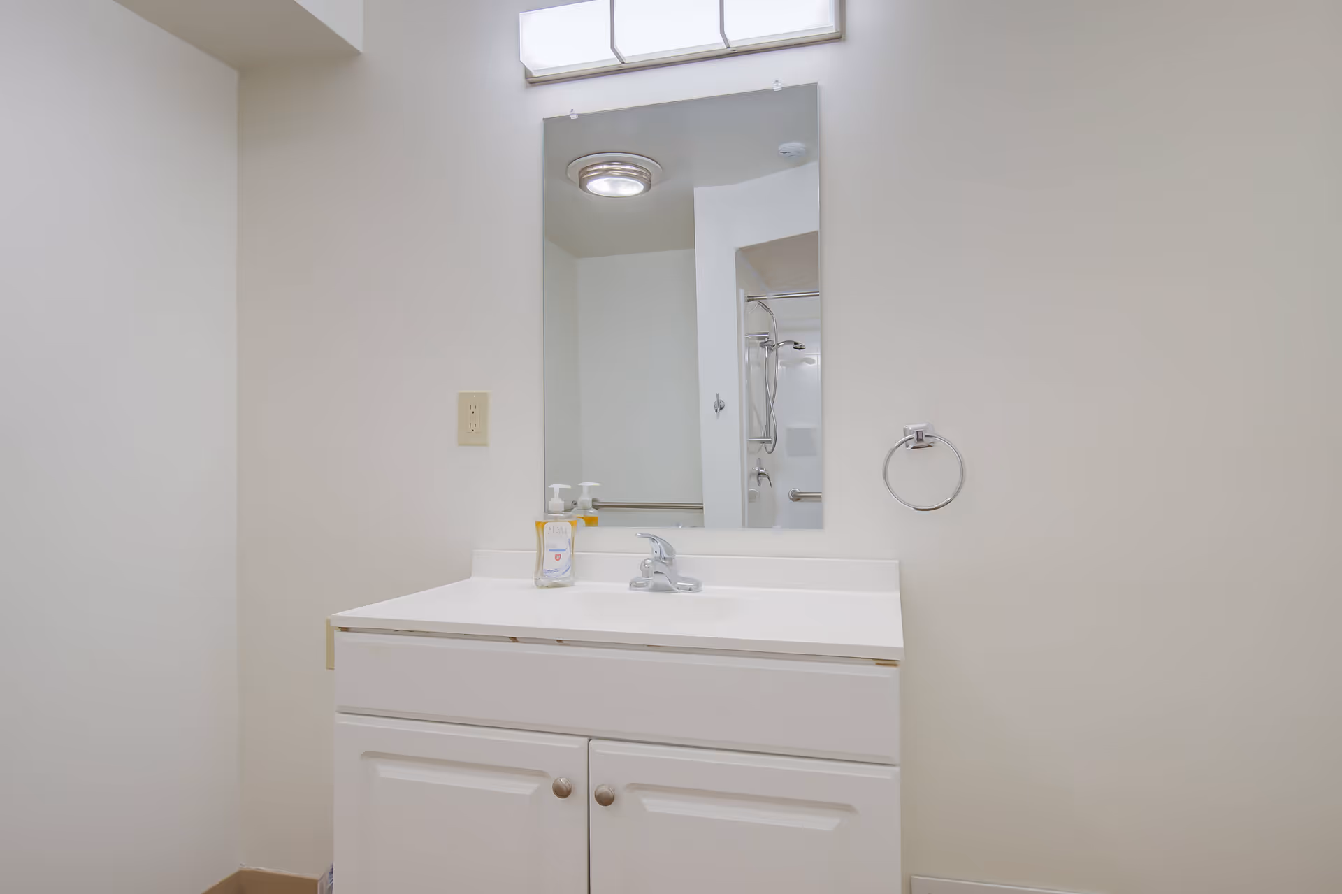 A clean bathroom vanity with a white countertop and cabinet, a faucet, a soap dispenser, and a mirror above. The mirror reflects a shower area with a grab bar and showerhead. A towel ring is mounted on the wall to the right of the vanity.