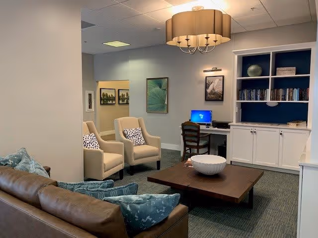 A cozy living room area in Lake Howard Heights featuring a brown leather sofa with blue patterned pillows, two beige armchairs with patterned cushions, a wooden coffee table with a decorative bowl, a built-in white cabinet with shelves holding books and decor, a desk with a computer and chair, and wall art. The room is softly lit by a ceiling light fixture.
