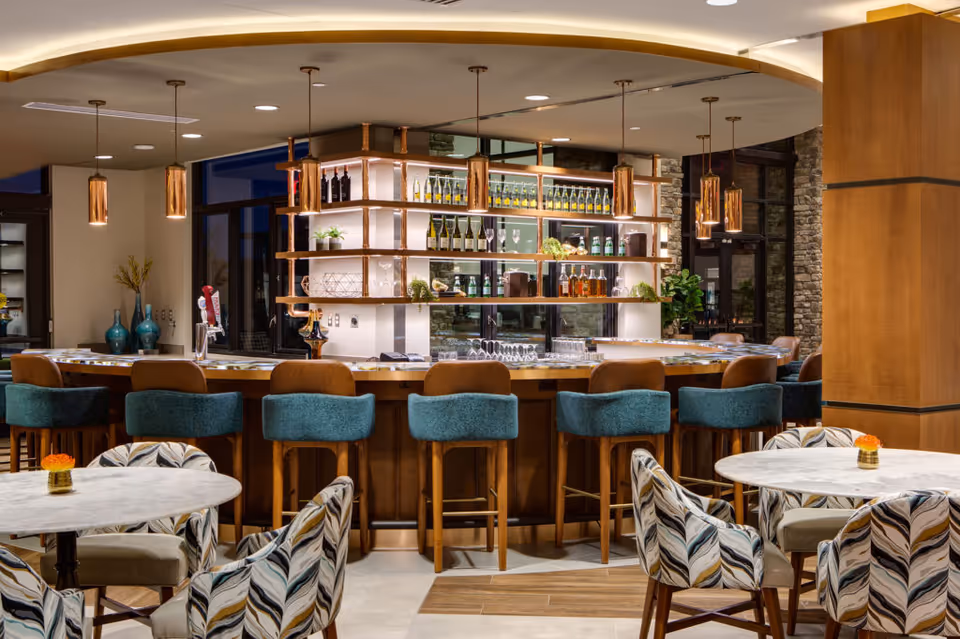 A modern bar area with a curved wooden counter surrounded by teal and brown bar stools. Behind the bar, shelves display various bottles of wine and liquor, glassware, and decorative plants. In the foreground, there are round tables with patterned upholstered chairs and small orange flower centerpieces. The space has warm lighting and a cozy, inviting atmosphere.
