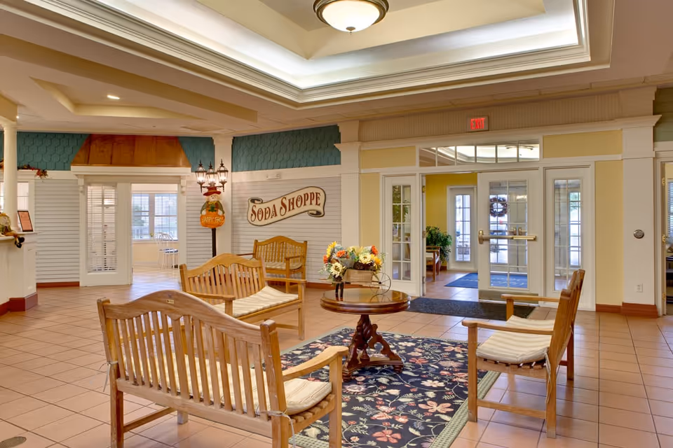 A spacious and well-lit common area with wooden benches and chairs arranged around a small round table with a floral centerpiece. The floor is tiled, and a decorative rug is placed under the table. The walls are painted in soft yellow and white tones with a sign that reads 'Soda Shoppe'. There are glass doors leading to another room and an exit door with windows allowing natural light to enter.