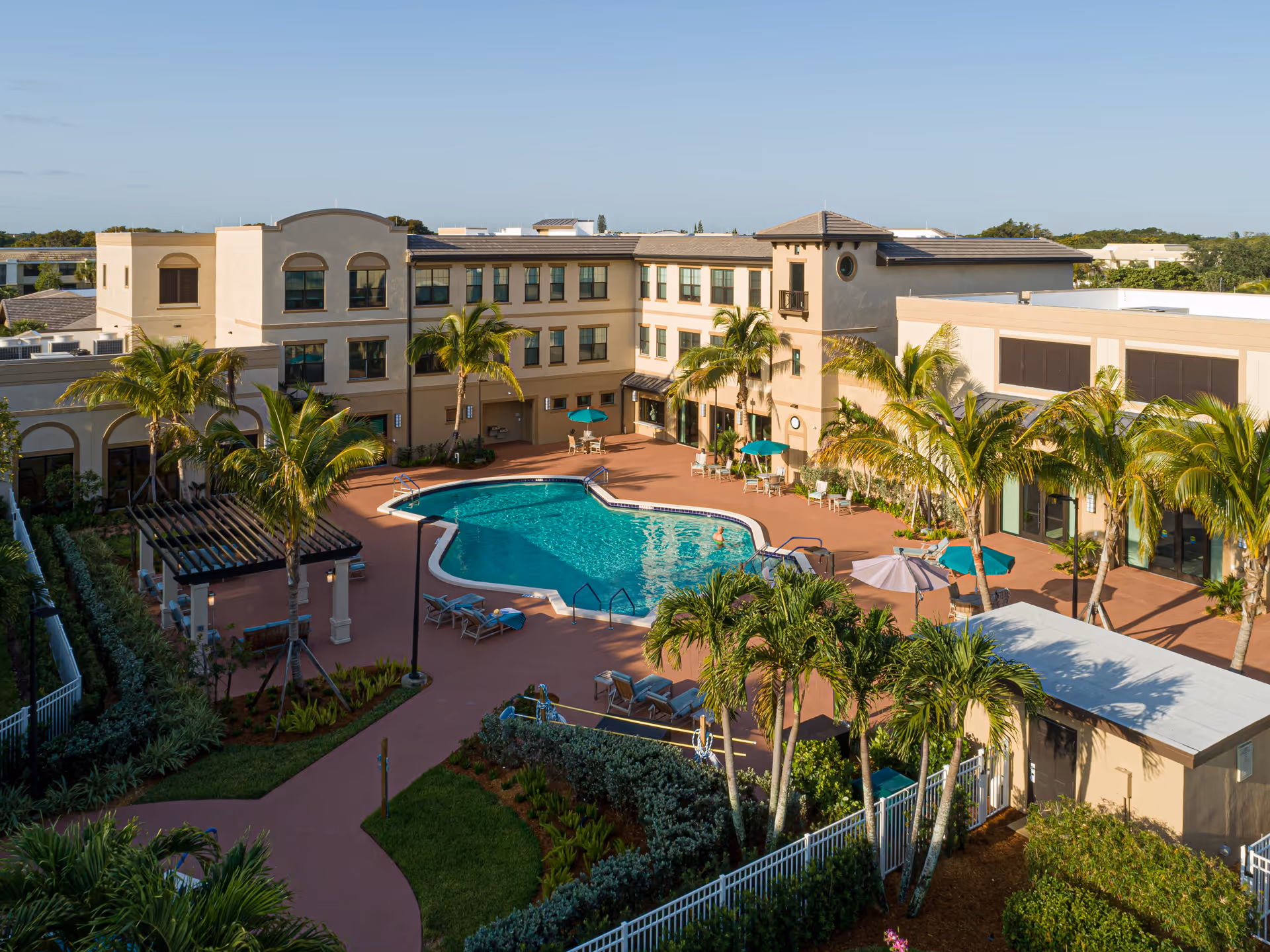 Outdoor view of Cascades at Delray Beach senior living facility featuring a large swimming pool surrounded by lounge chairs and umbrellas, palm trees, and a three-story beige building under a clear blue sky.