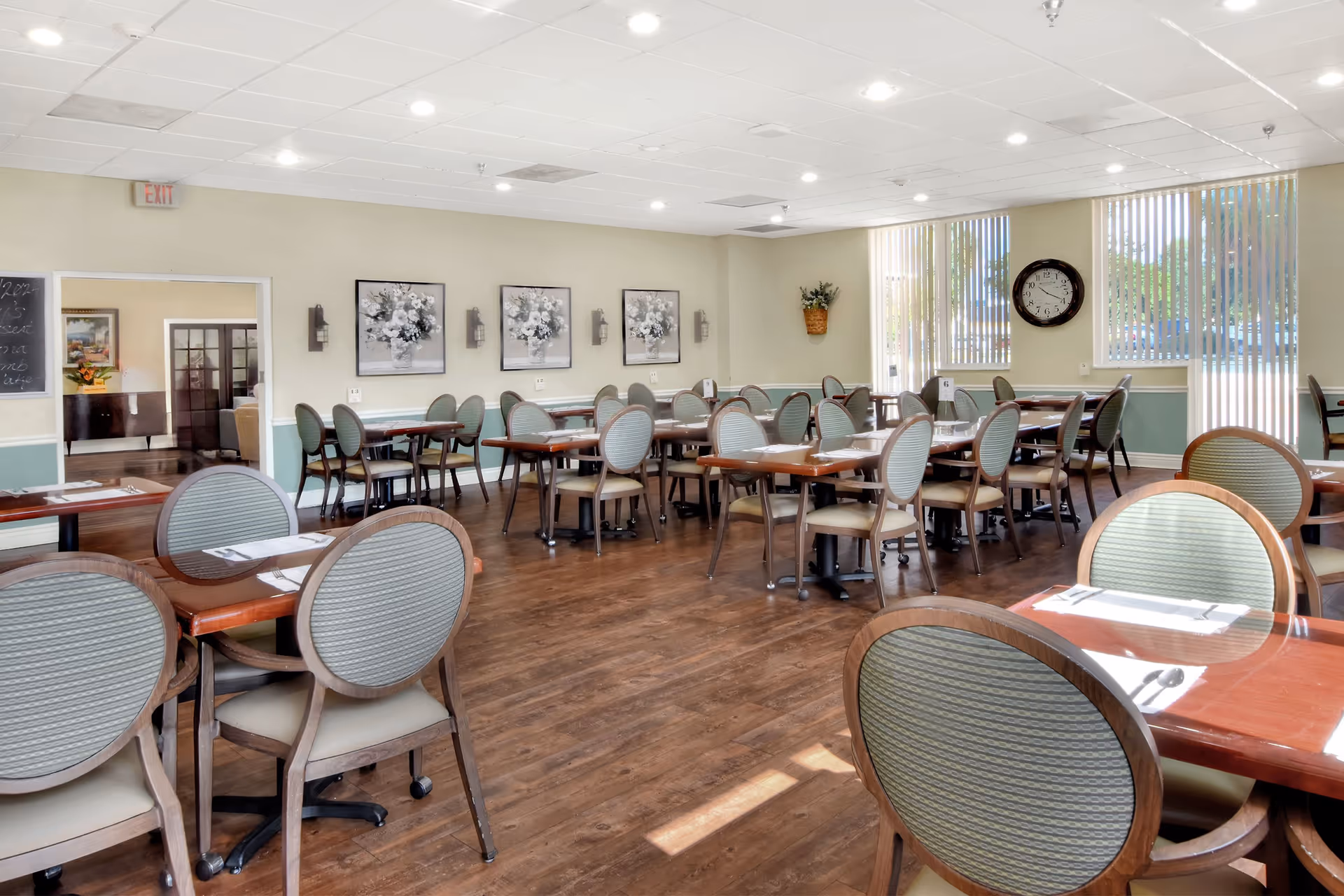 Bright communal dining room with multiple tables and round-backed chairs, large windows, and wood flooring.