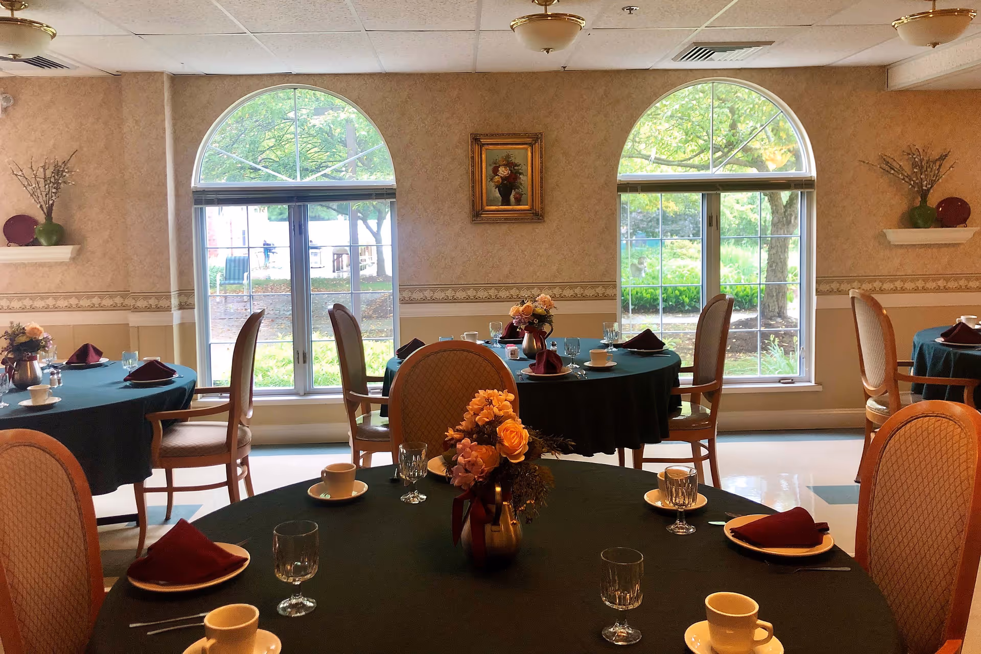 Dining room with round tables covered in dark green tablecloths, each set with cups, glasses, plates, and burgundy napkins. Floral centerpieces decorate the tables. The room has large arched windows showing greenery outside, beige walls with a decorative border, and framed artwork between the windows.