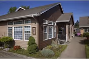 Front exterior of a single-story brown residence with large multi-pane windows, a sidewalk entry, and landscaped shrubs.