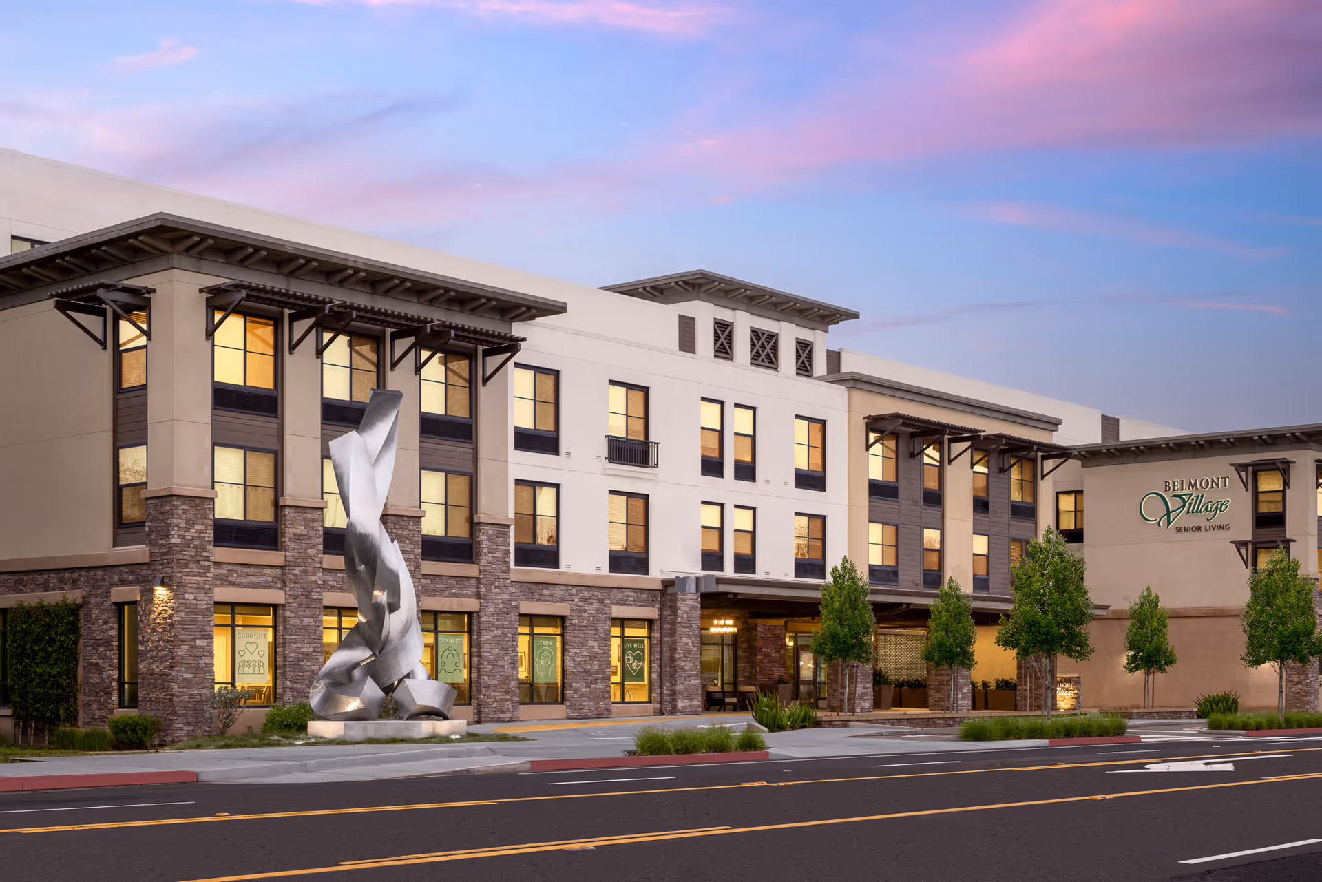 Exterior front of a three-story Belmont Village Senior Living building with illuminated windows, a modern metal sculpture, and landscaped entrance at dusk.