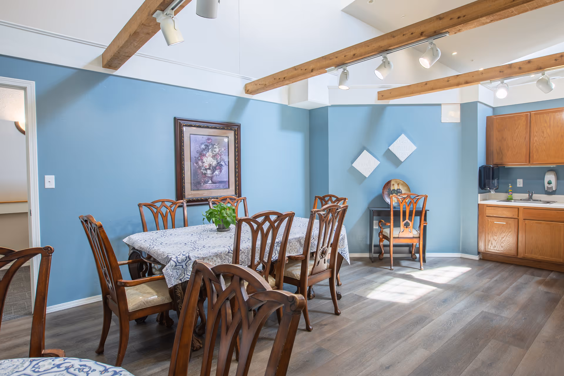 A dining area with a wooden table covered by a patterned tablecloth and surrounded by wooden chairs. The walls are painted blue with decorative artwork hanging. There is a small kitchenette with wooden cabinets, a sink, and soap dispensers. The ceiling has exposed wooden beams with track lighting.