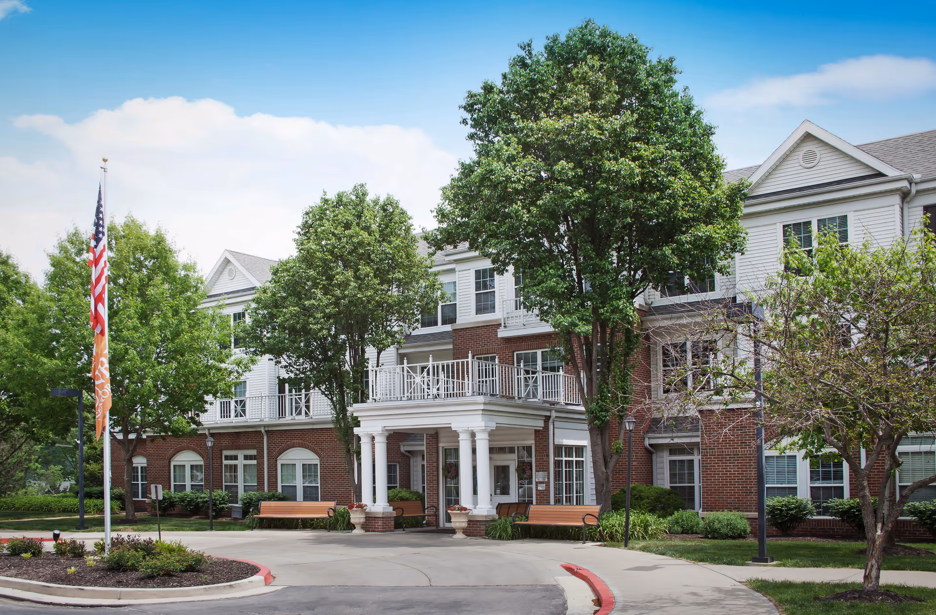 Front exterior view of a senior living facility named The Village at Mission, showing a brick and white-paneled building with large windows, a covered entrance with white columns, benches, trees, and an American flag on a flagpole.