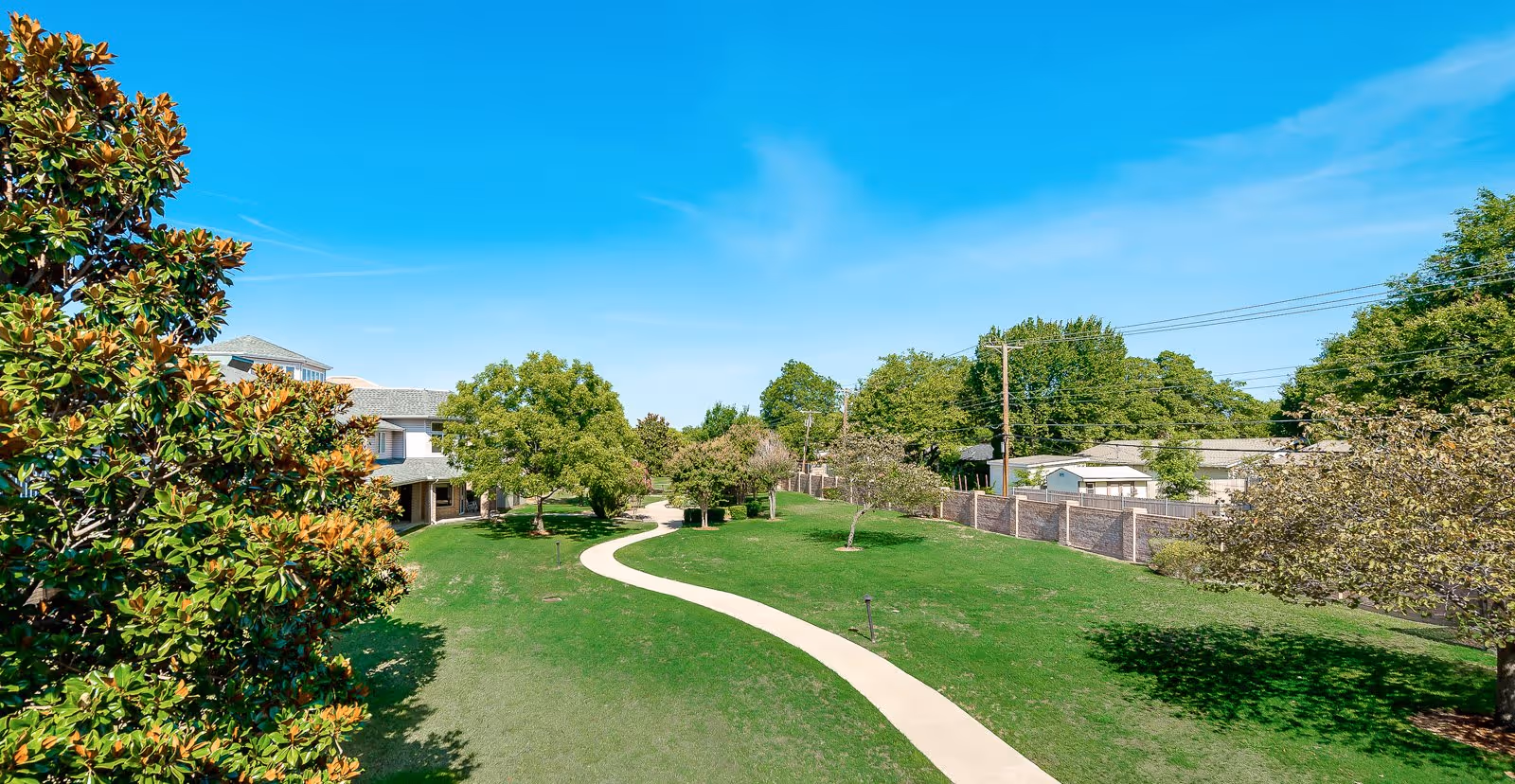 A bright outdoor scene showing a winding concrete pathway through a well-maintained grassy area with various trees and shrubs. In the background, there are buildings partially visible behind the trees under a clear blue sky.
