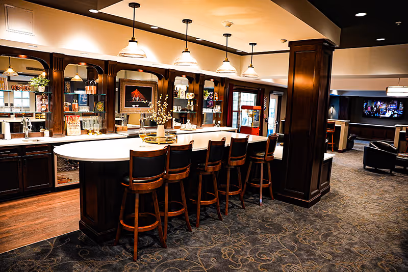 Warmly lit communal bar and lounge with a long curved counter, five wooden bar stools, pendant lights, and dark wood cabinetry.