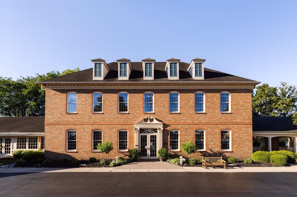 Front exterior view of a two-story brick building with multiple windows and a central entrance door, surrounded by small shrubs and a bench on the right side, under a clear blue sky.