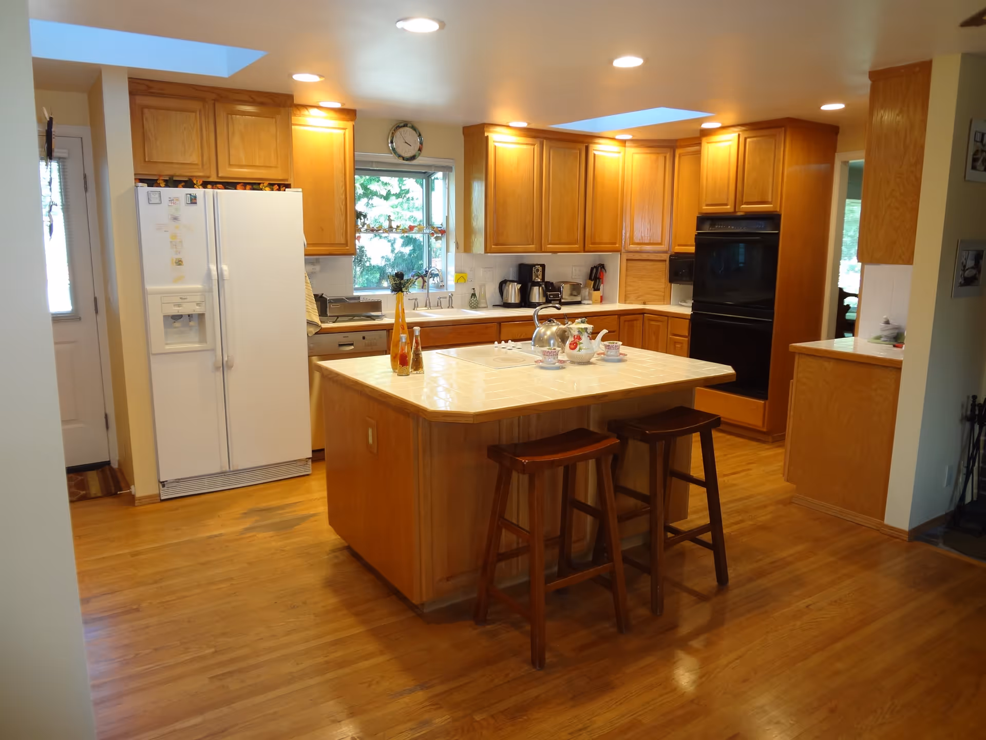A spacious kitchen with wooden cabinets, a white refrigerator, a built-in oven, and a central island with a tiled countertop. Two wooden stools are placed at the island, which has a tea set and decorative bottles on it. The kitchen has hardwood floors and recessed lighting, with a window above the sink letting in natural light.