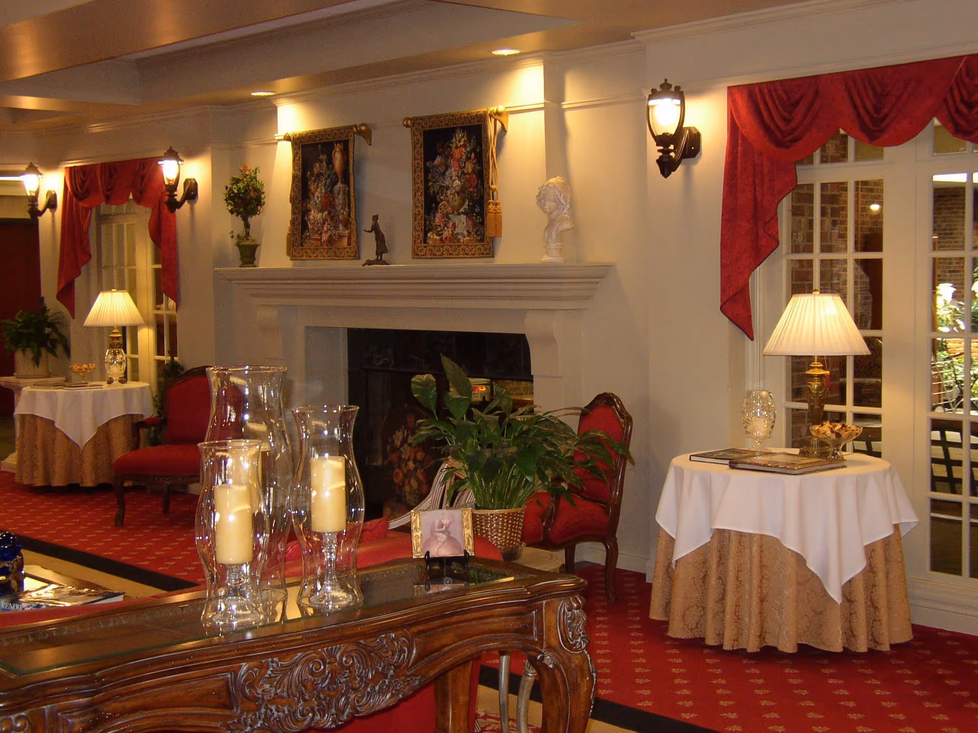 A cozy and elegantly decorated living room area with a fireplace, red carpet, and classic furniture. There are two red chairs near the fireplace, a wooden table with glass candle holders in the foreground, and round tables with white and beige tablecloths, lamps, and decorative items. The walls are adorned with framed floral artwork and a white bust sculpture. Red drapes frame the windows and doors, adding a warm and inviting atmosphere.