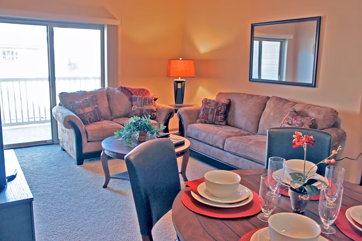A cozy living room area with two beige sofas adorned with patterned cushions, a wooden coffee table with a green plant centerpiece, a side table with a lamp, and a large mirror on the wall. In the foreground, there is a dining table set with white bowls, plates, glasses, red placemats, and a small potted orchid. Sliding glass doors lead to an outdoor balcony.