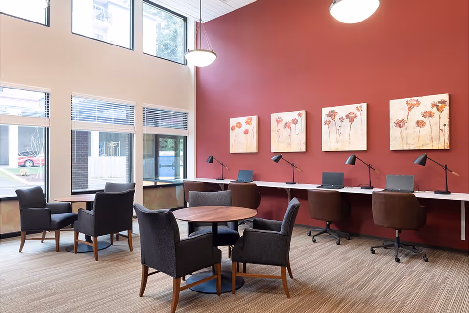 Bright communal lounge with round tables and armchairs and a row of workstations with laptops and lamps against a red accent wall.