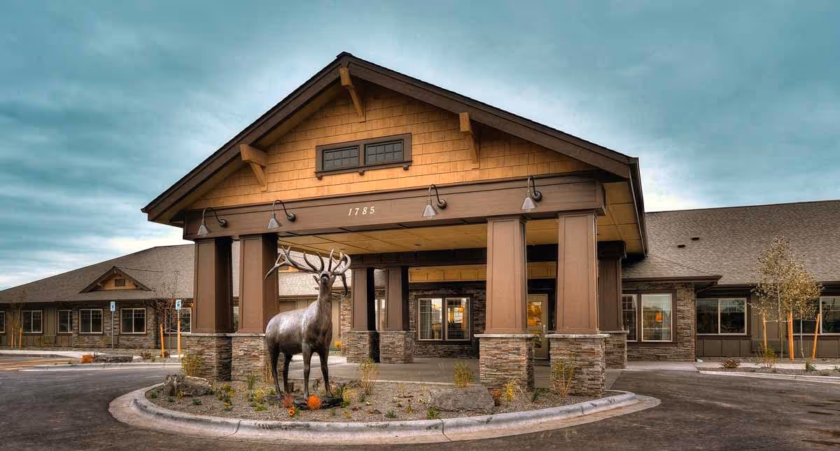 Exterior view of a single-story building with a covered entrance supported by large pillars. In front of the entrance, there is a statue of a deer standing on a landscaped circular island. The building has a rustic design with stone and wood elements, and the address number 1785 is displayed above the entrance.