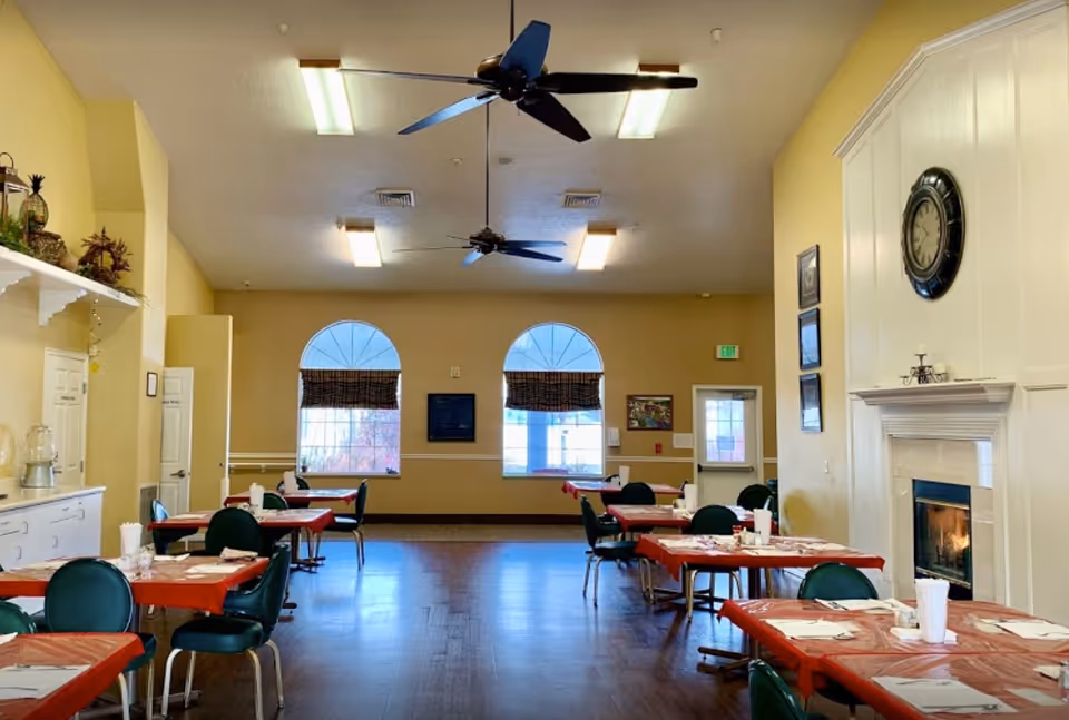 Spacious dining room with several tables covered in red tablecloths, green chairs, ceiling fans, arched windows and a fireplace.