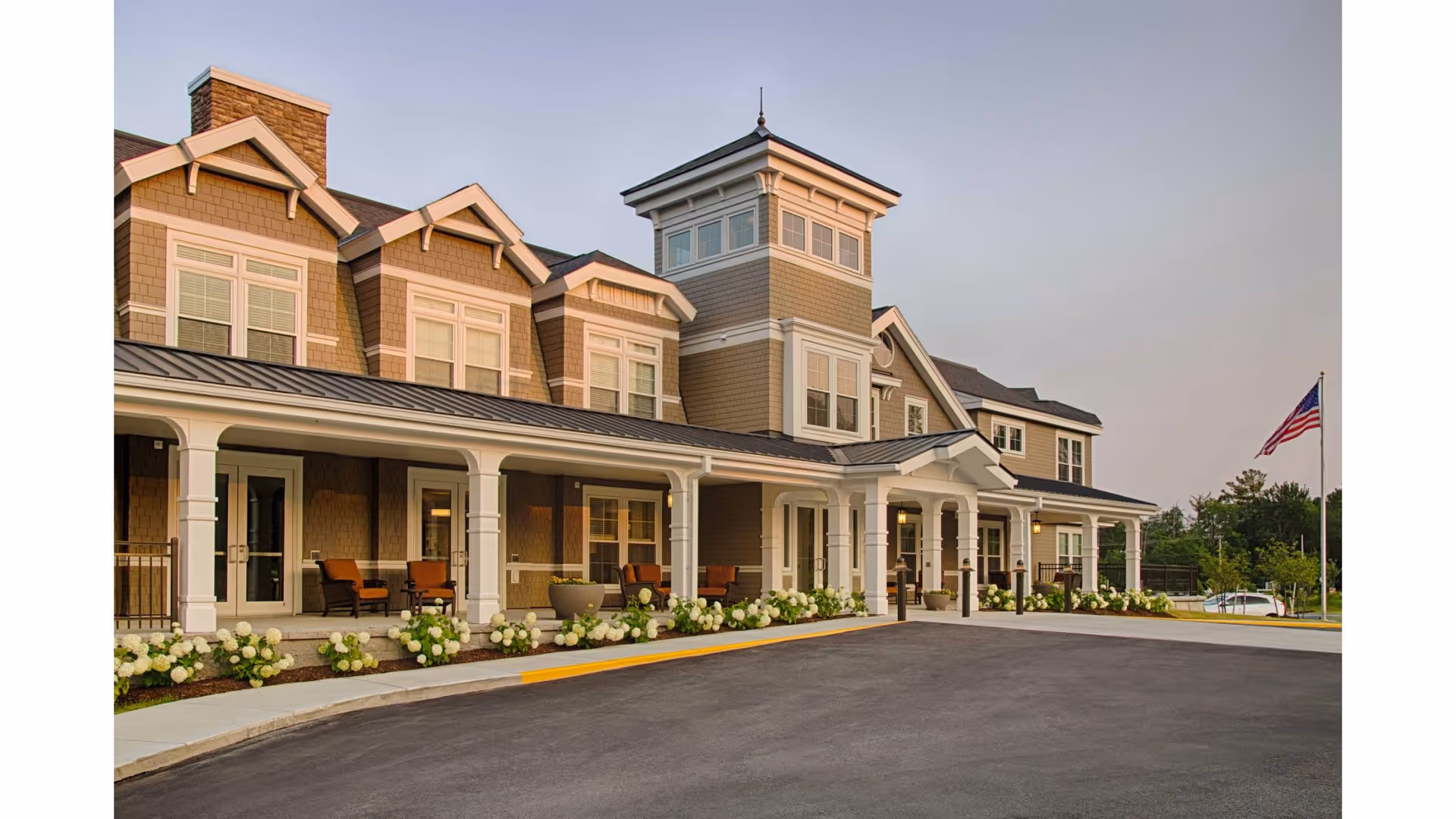 Exterior view of a large, two-story senior living facility building with a covered porch, several windows, outdoor seating, flower beds, and an American flag on a flagpole in the background during early evening.