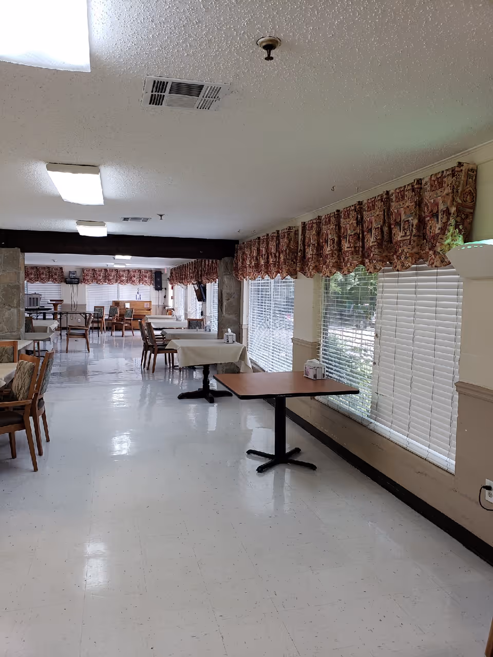 Long dining room with tables and chairs, large windows with patterned valances along the right wall and a tiled floor.