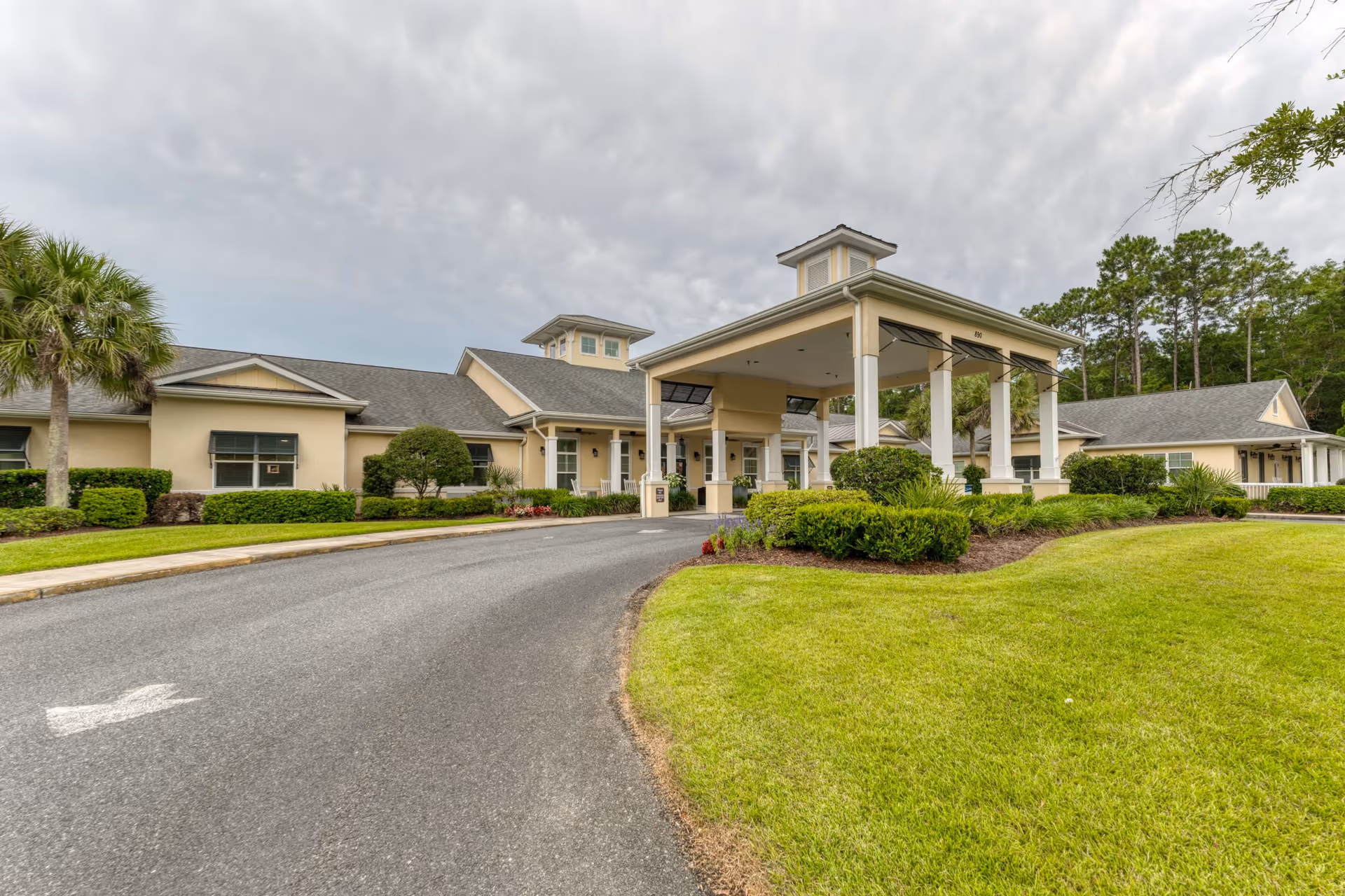 Exterior view of Addington Place of Brunswick, showing a single-story building with a covered entrance driveway, surrounded by well-maintained green lawns, bushes, and trees under a cloudy sky.