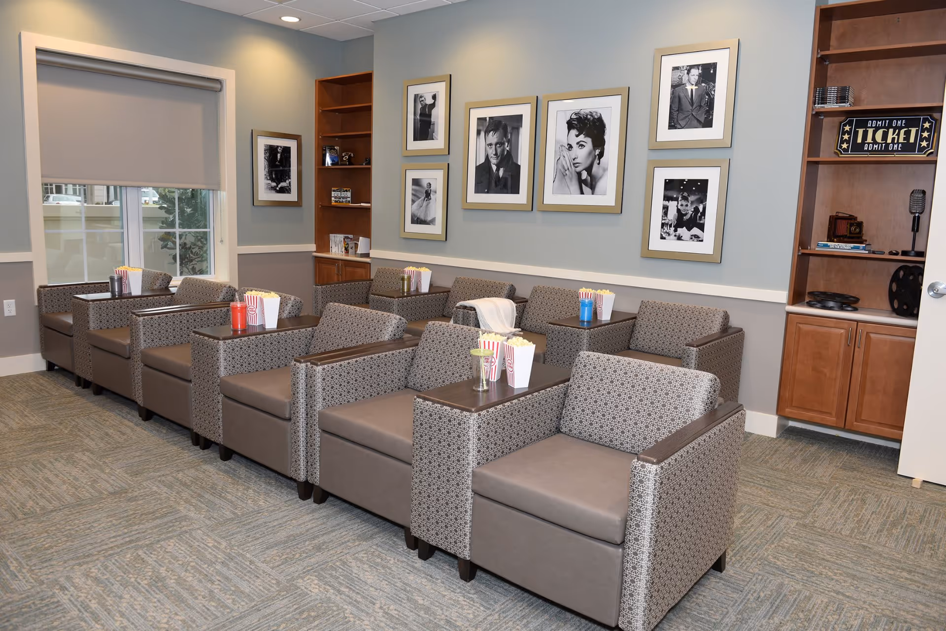 A cozy senior living common area with multiple patterned armchairs arranged in rows, each with a small table holding popcorn containers and drinks. The walls are decorated with framed black-and-white photos of classic movie stars. There are built-in wooden shelves with vintage decor items and a window with a roller shade.