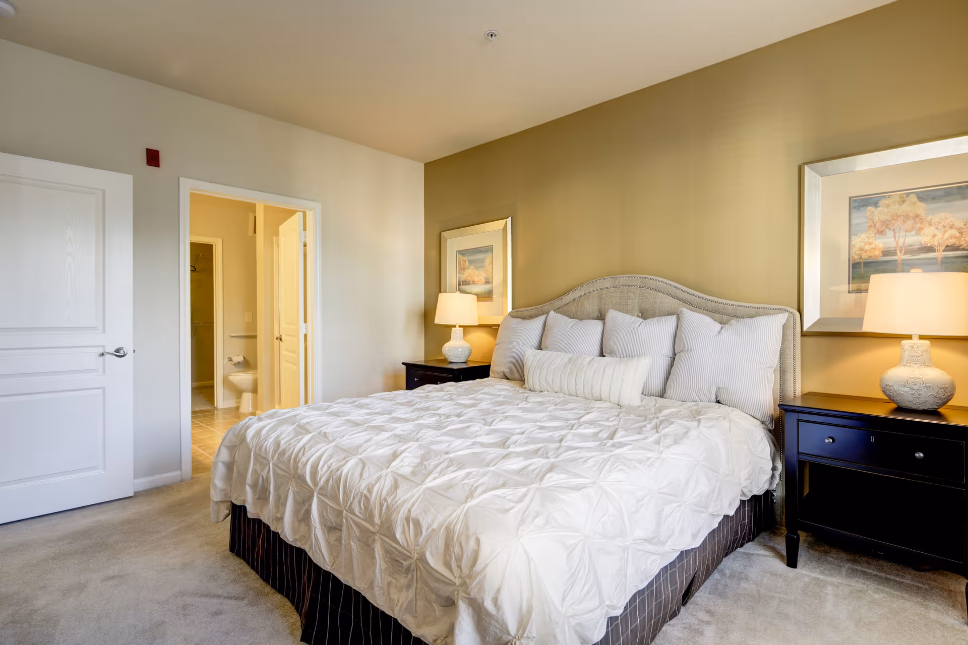 A neatly made bed with white bedding and multiple pillows in a bedroom. The room has beige walls, two black nightstands with matching lamps, and framed artwork above each nightstand. An open door reveals a bathroom with a toilet and tiled floor.
