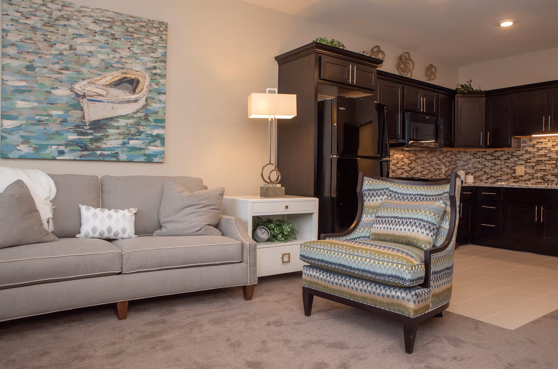 Open living area showing a gray sofa, patterned armchair, side table with a lamp, and a kitchenette with dark wood cabinets in the background.