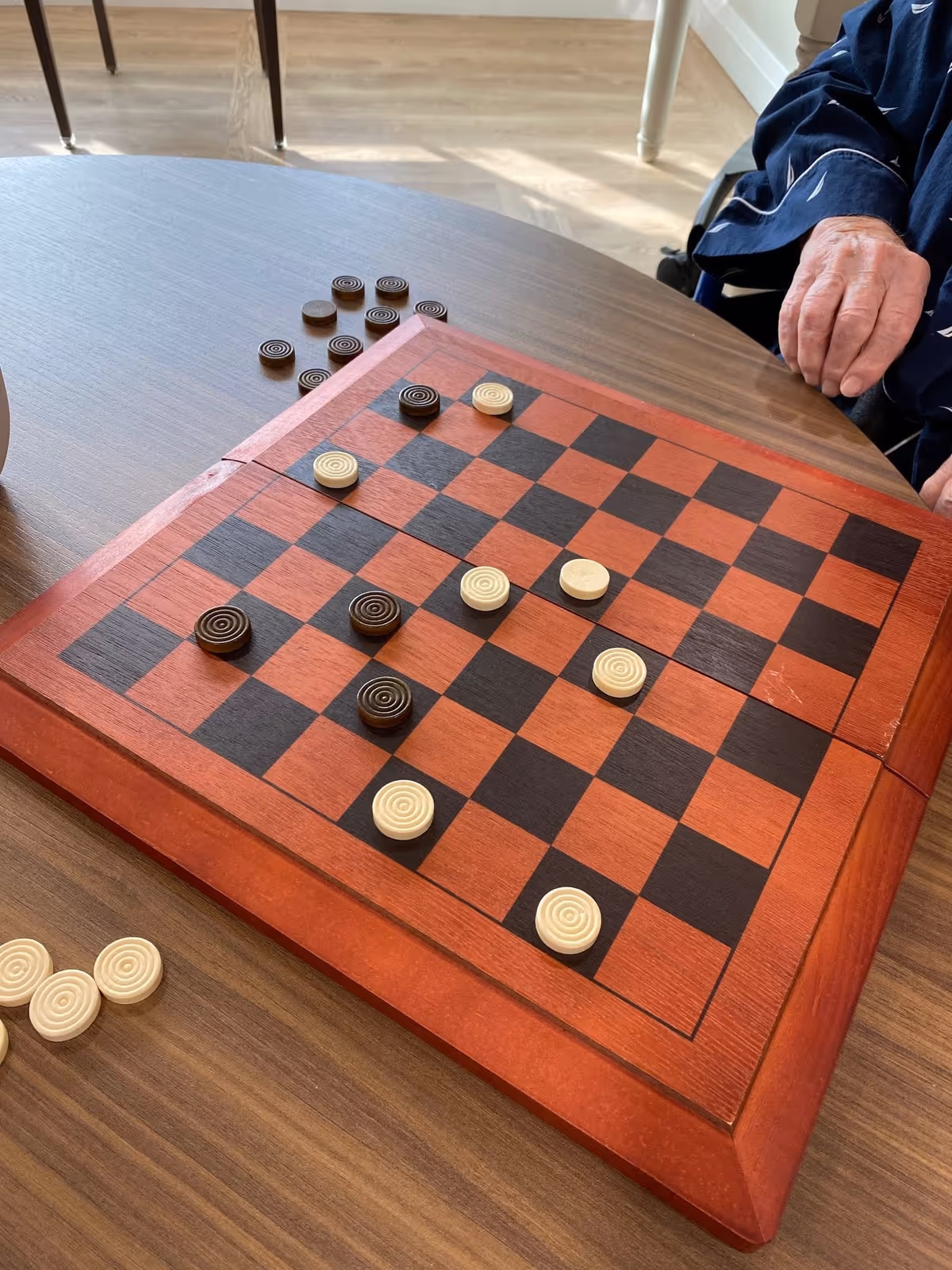 A close-up view of a wooden checkers board on a table with black and white checkers pieces arranged for a game. A person's hand is visible on the right side, resting near the board. The setting appears to be indoors with natural light coming in.