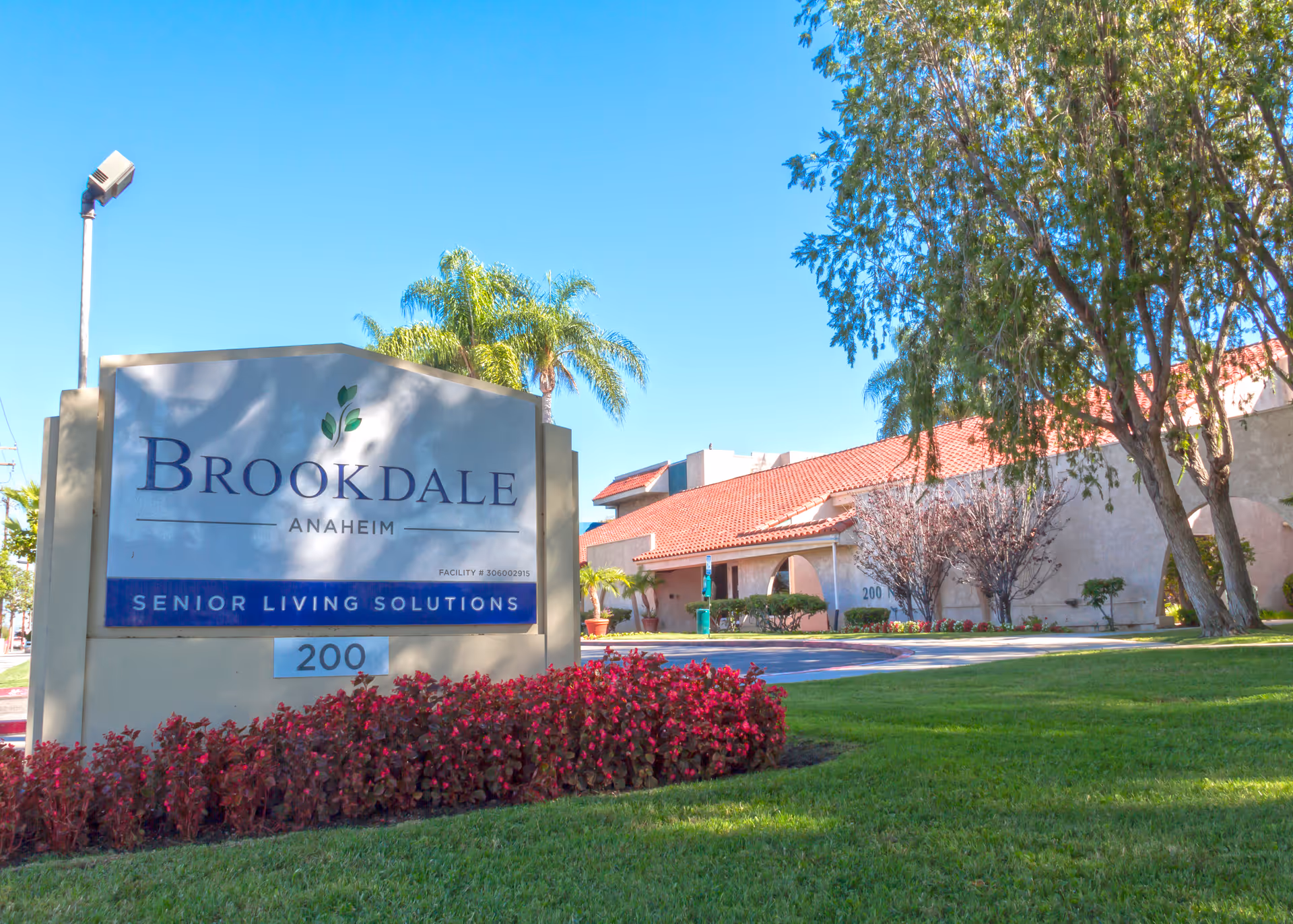 Exterior view of Brookdale Anaheim senior living facility with a large sign in the foreground that reads 'Brookdale Anaheim Senior Living Solutions' and the number 200. The building has a red-tiled roof and is surrounded by green grass, trees, and red flowering plants under a clear blue sky.