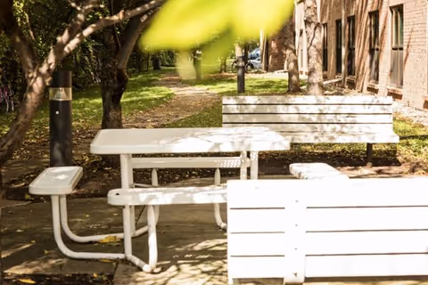 White picnic table and benches in a shaded courtyard next to a brick building and trees.