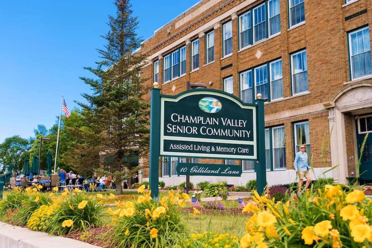 Outdoor view of Champlain Valley Senior Community building with a green sign in front that reads 'Champlain Valley Senior Community Assisted Living & Memory Care 10 Gilliland Lane'. There are yellow flowers and greenery in the foreground, people sitting at tables with umbrellas on the left side, and a person walking near the building entrance on the right side.