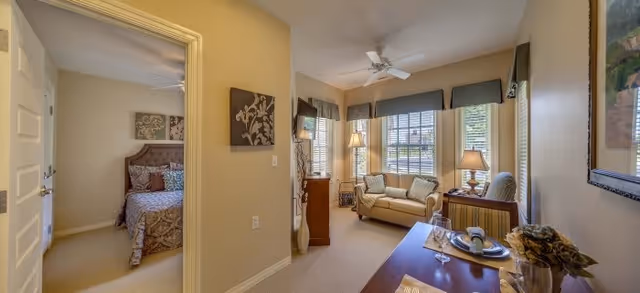 Interior view of a senior living facility showing a cozy living room with a beige sofa, armchair, side table with lamp, and a ceiling fan. Adjacent to the living room is a bedroom with a bed covered in patterned bedding. The walls are painted light beige and decorated with framed artwork.