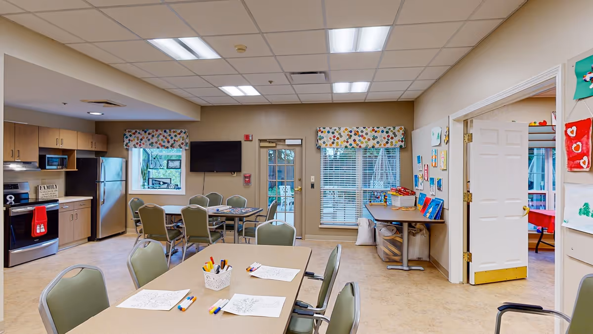 Bright communal activity and dining room with a kitchenette, multiple tables and chairs, and arts-and-crafts supplies.