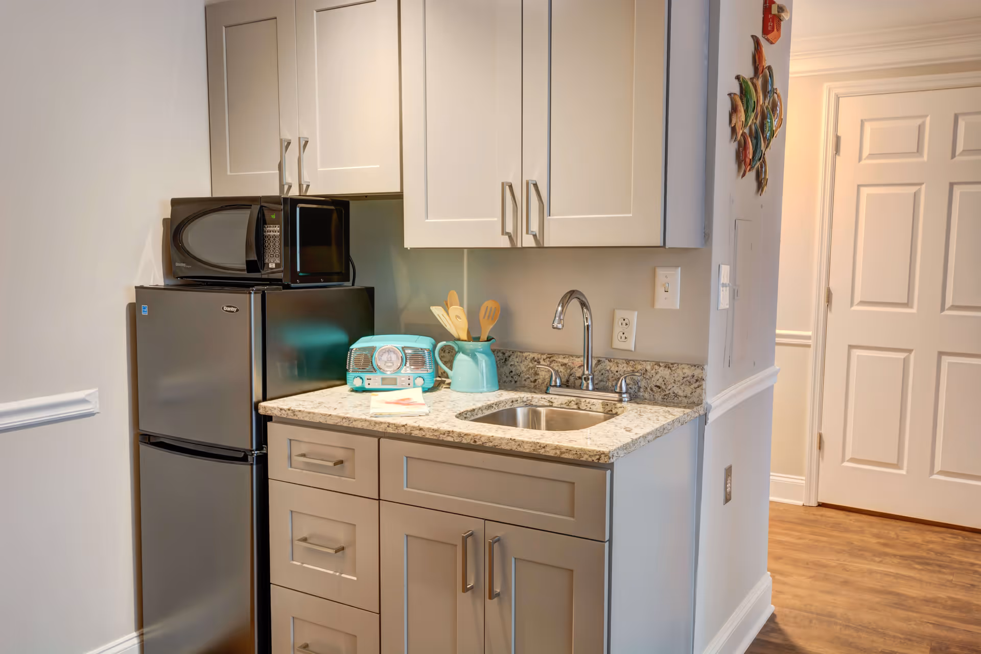 A small kitchenette area with a granite countertop, a stainless steel sink, white upper cabinets, and gray lower cabinets. On the countertop, there is a turquoise retro-style radio, a matching turquoise pitcher holding wooden utensils, and a folded napkin. A black microwave sits on top of a compact black refrigerator. The floor is wooden, and a white door is visible in the background.
