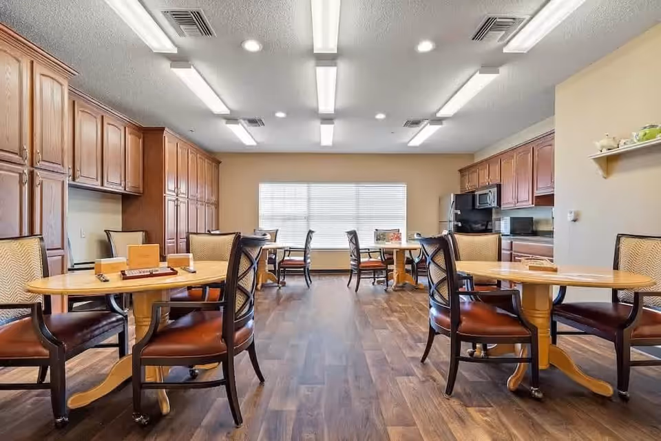 A bright dining room with wooden tables and cushioned chairs arranged neatly on a wood floor. The room features large wooden cabinets along the walls, a window with blinds letting in natural light, and a kitchen area with a microwave and refrigerator in the background.