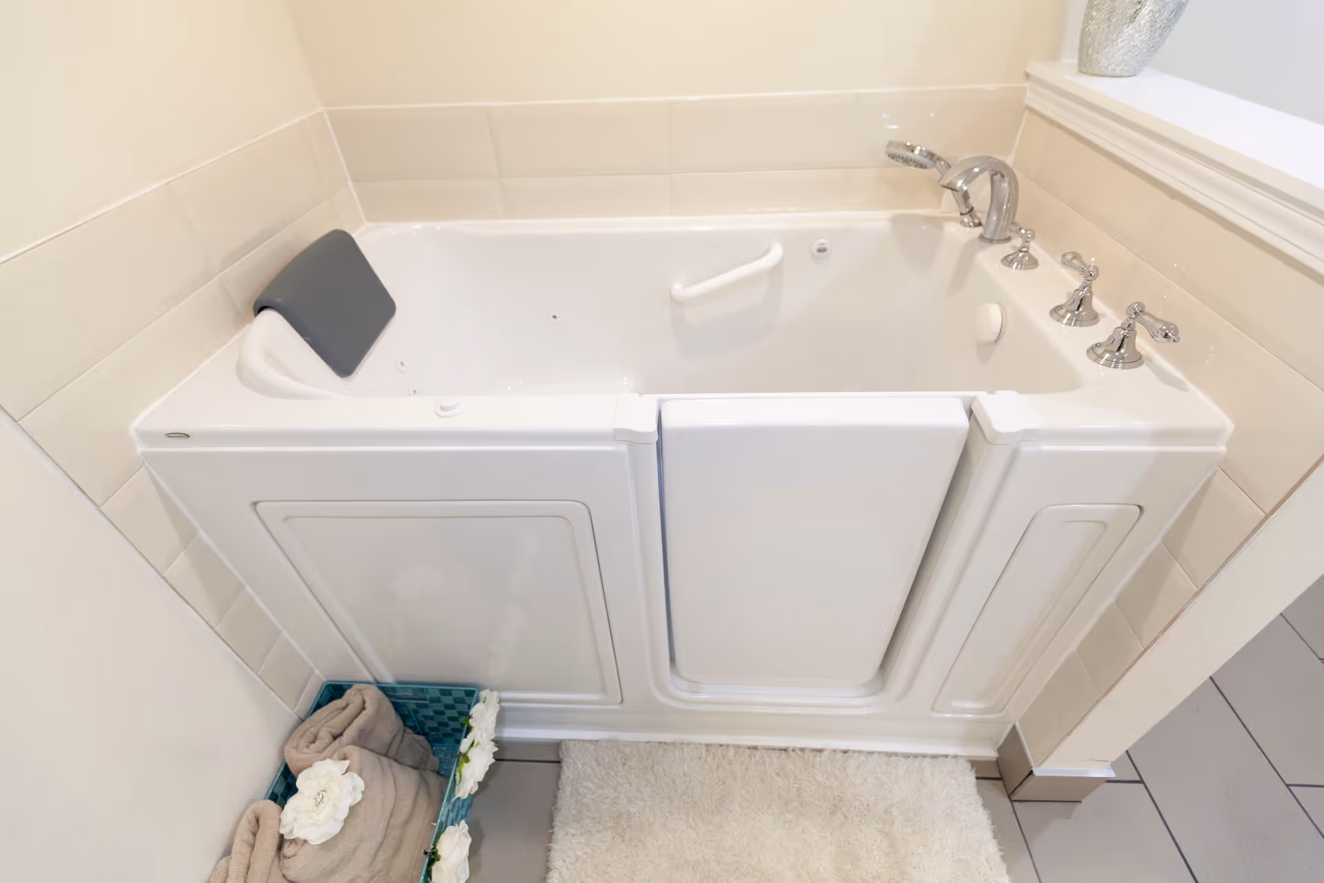 Walk-in bathtub with a faucet, grab bar and headrest in a tiled bathroom next to a towel basket and bath mat.
