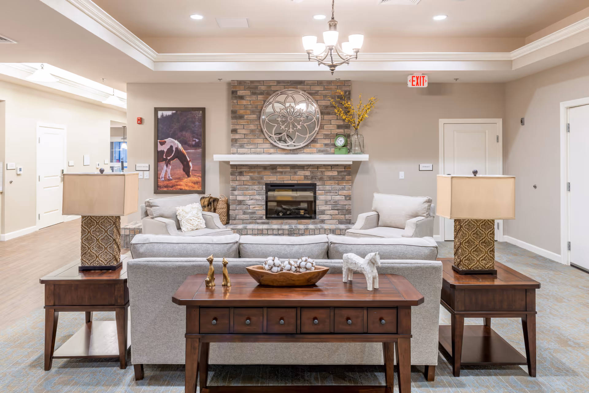 A cozy senior living facility common area with a brick fireplace centered on the back wall. In front of the fireplace are two light gray armchairs and a matching sofa. A wooden console table with decorative items, including two small giraffe figurines and a white horse figurine, is placed behind the sofa. Two matching wooden side tables with patterned lamps flank the sofa. A framed picture of a horse grazing is on the left wall, and a round decorative mirror hangs above the fireplace. The room has beige walls, a chandelier, and an exit sign above a door on the right.