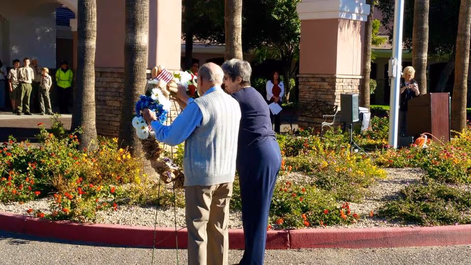 An elderly man and woman stand outdoors near a landscaped area with flowers and palm trees, placing a decorative wreath. In the background, a group of people, including some in uniform, stand near a building entrance.