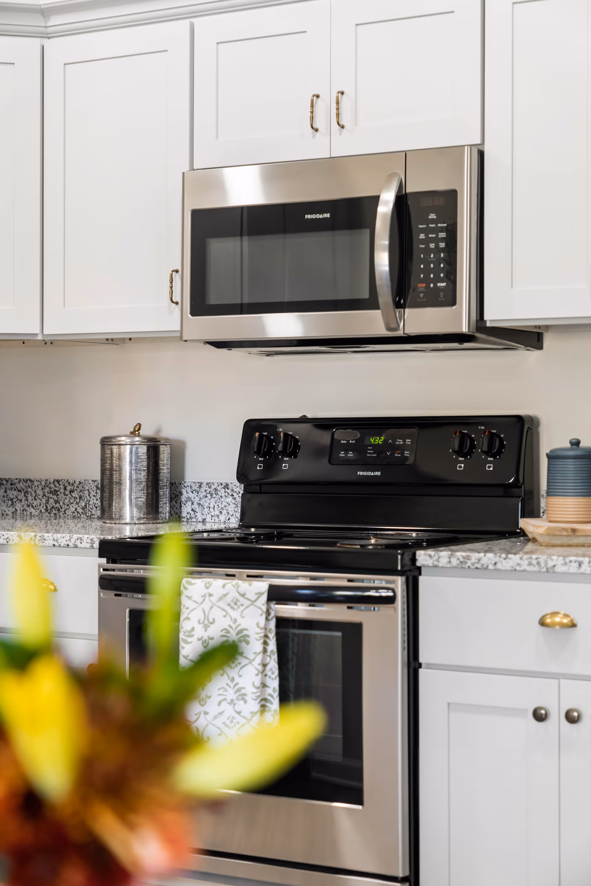 Stainless steel microwave and oven set into white kitchen cabinets above a granite countertop with a utensil canister and a patterned towel on the oven door.